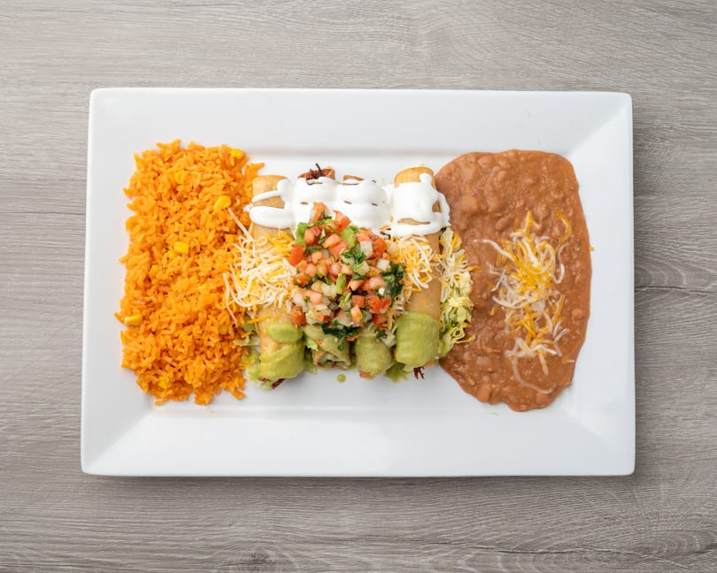 A white rectangular plate holds four taquitos topped with sour cream, pico de gallo, and guacamole, alongside orange rice and refried beans.