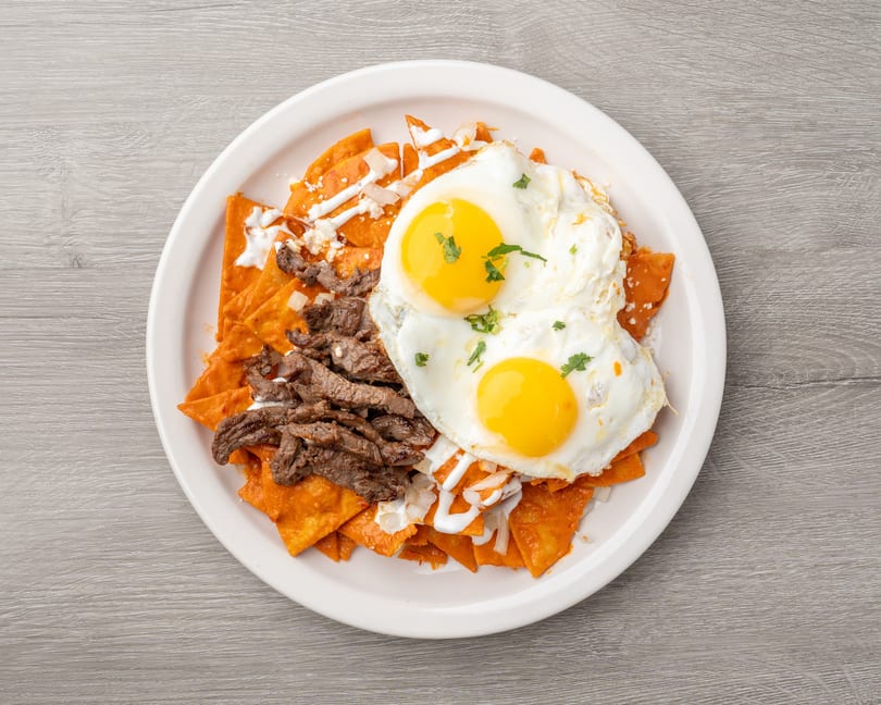 Chilaquiles with red sauce, steak, two fried eggs, sour cream, and cheese on a white plate.