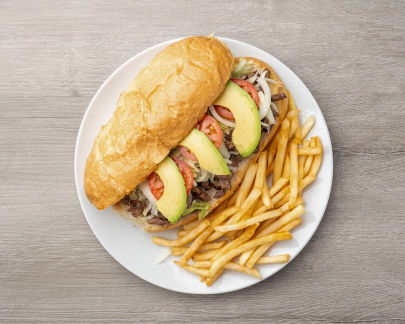 Steak sandwich with avocado, tomato, onion, and lettuce, served with french fries on a white plate.