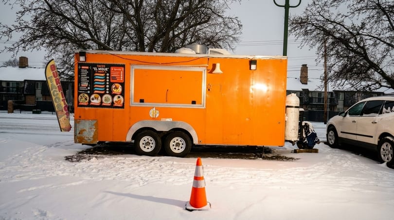 An orange food truck, "Tacos El Mibue," sits in a snowy parking lot with a white car nearby.