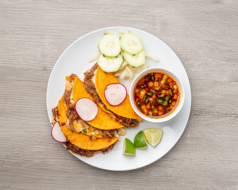 Birria tacos with radishes, cucumbers, limes, and a side of consommé on a white plate.