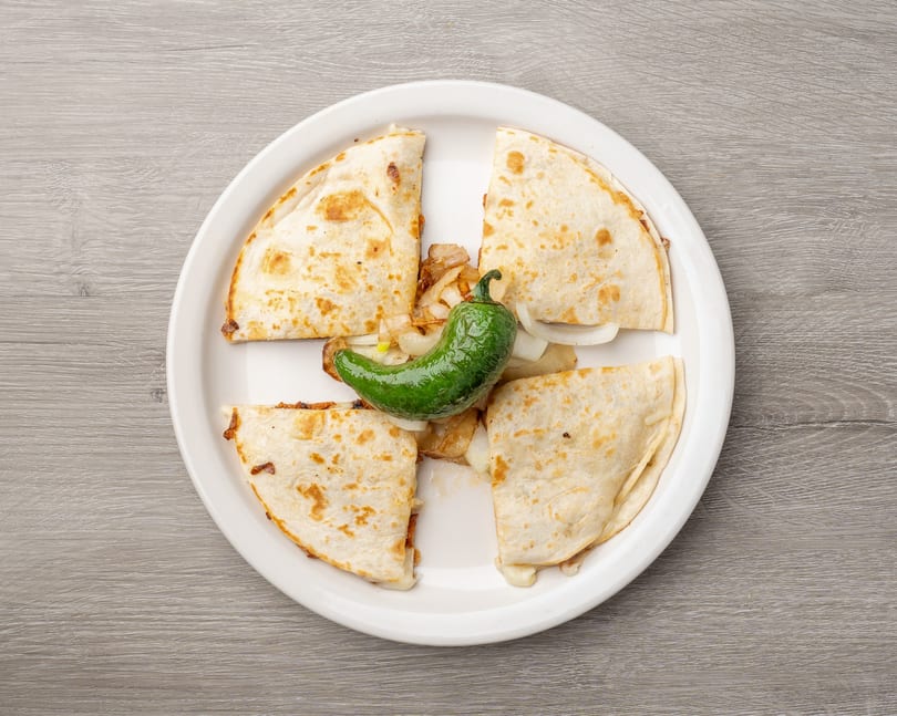 Four quesadilla wedges with melted cheese, grilled onions, and a green chili pepper on a white plate.