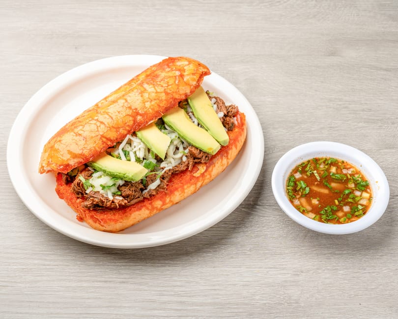 A Mexican torta with shredded meat, avocado, and cheese, served with a side of consommé.