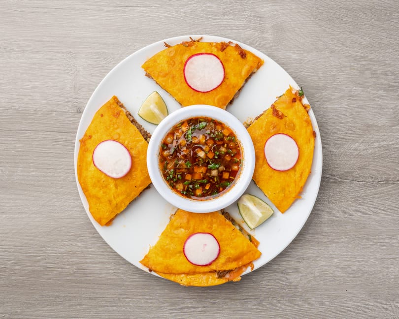 Four golden-brown quesadillas with radish slices and lime wedges surround a bowl of dipping sauce.