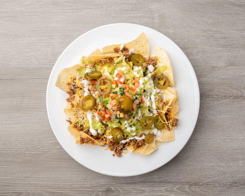 A white plate filled with nachos, topped with ground beef, cheese, lettuce, pico de gallo, sour cream, guacamole, and jalapeños.